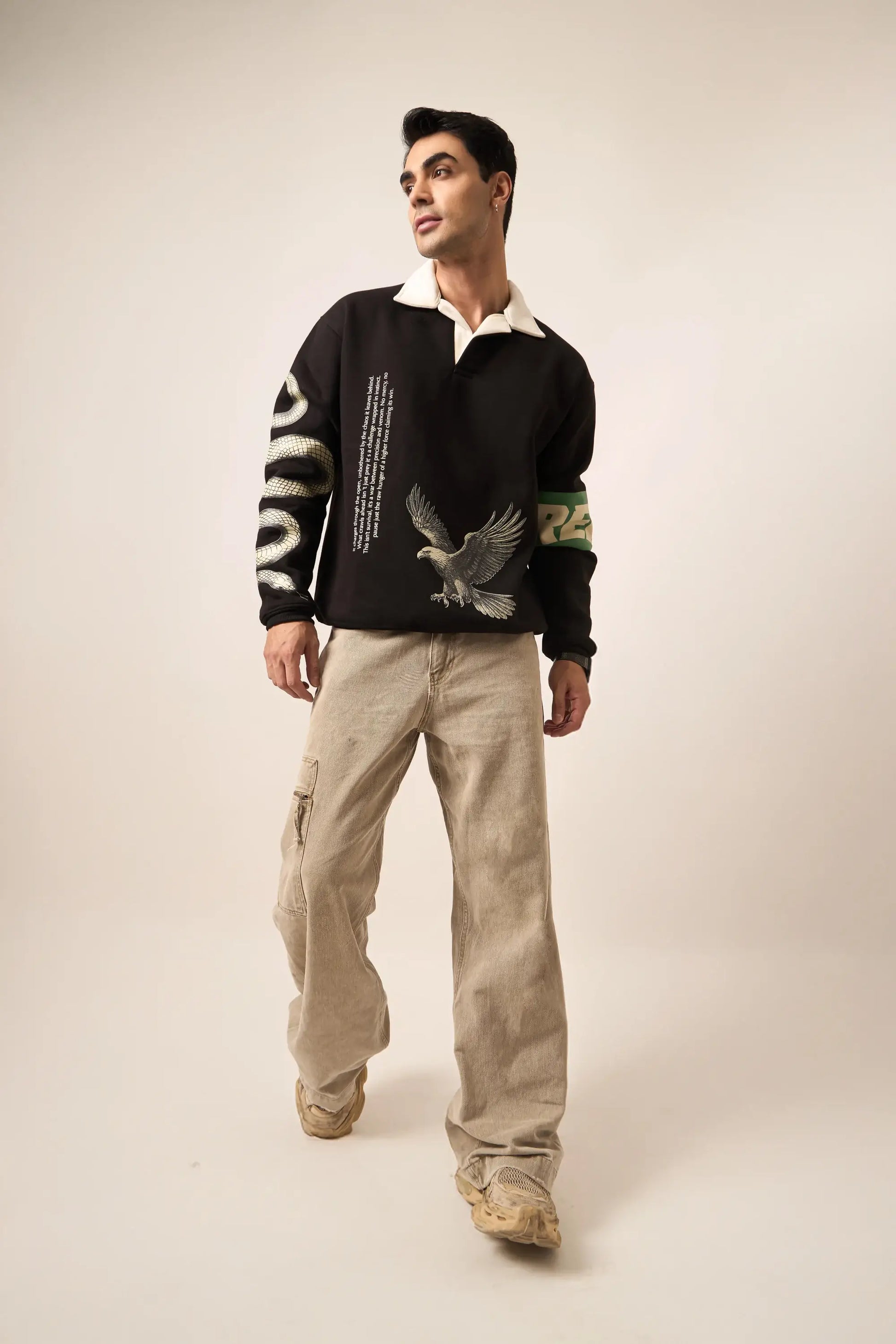 Man wearing a black polo sweatshirt with bird and serpent designs and beige pants on a plain background